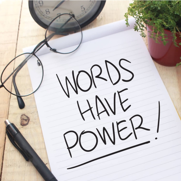 Notepad on a wooden desk with 'Words Have Power!' handwritten in bold letters, surrounded by glasses, a pen, a clock, and a potted plant
