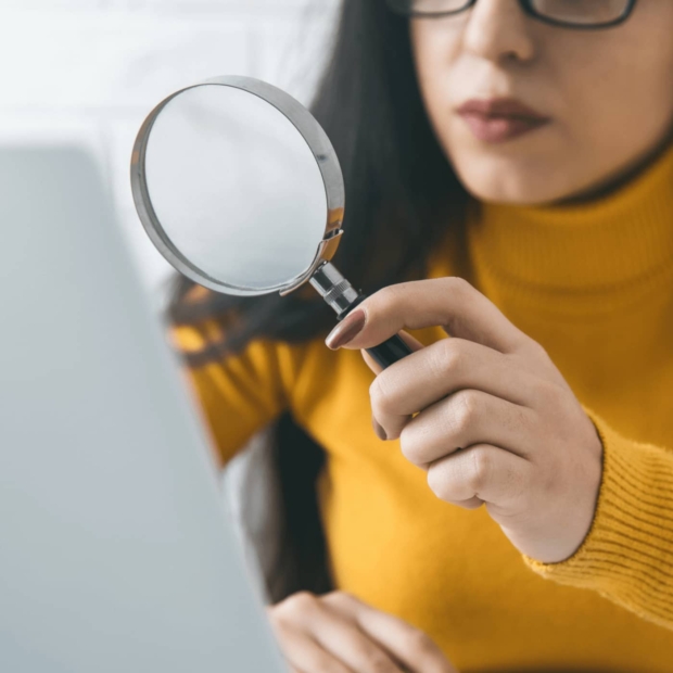 Close-up of a woman in a yellow jumper holding a magnifying glass up to a laptop screen, symbolising a digital audit.