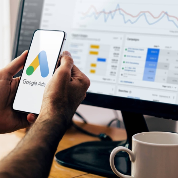Person holding a phone showing the Google Ads logo, with a campaign performance dashboard visible on a computer monitor in the background.