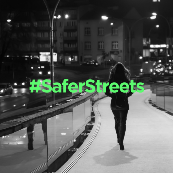 Black and white photo of a woman walking alone on a city street at night, overlaid with green '#SaferStreets.