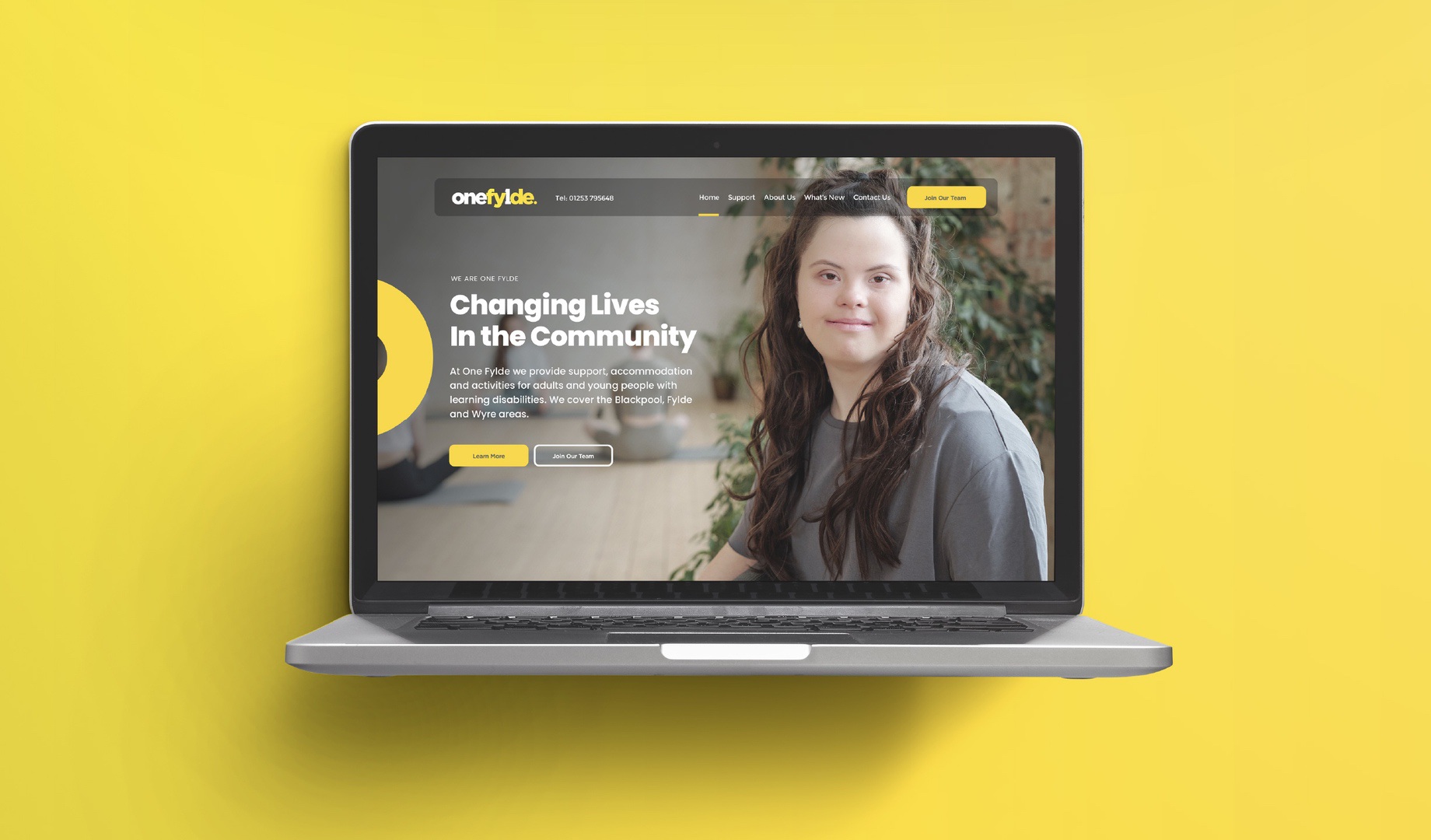 Laptop mockup of the One Fylde website homepage, showing the 'Changing Lives In the Community' banner and a smiling woman, on a yellow background.