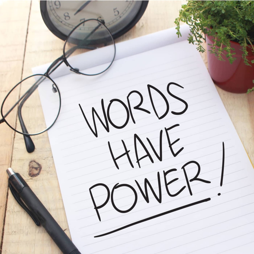Notepad on a wooden desk with 'Words Have Power!' handwritten in bold letters, surrounded by glasses, a pen, a clock, and a potted plant