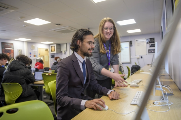 A staff member helping a student at a computer in a busy college IT classroom at Saint Francis Xavier Catholic Sixth Form College.