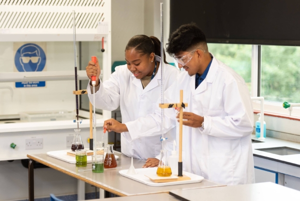 Two students in lab coats conducting a chemistry experiment with coloured liquids in beakers and flasks in a science lab at Saint Francis Xavier Catholic Sixth Form College.