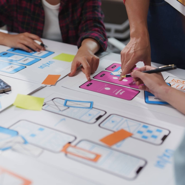 Close-up of a UX design team reviewing paper wireframes and app screen mockups spread across a table, with colourful sticky notes and sketches.